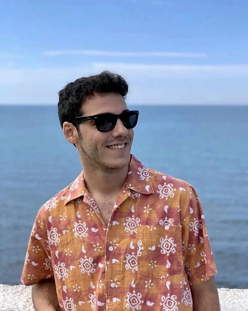 Un ragazzo sorridente davanti al mare, con occhiali da sole e una camicia a fantasia arancione.