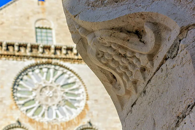Un capitello di pietra scolpita in primo piano; dietro, sfocato, il rosone di una chiesa romanica a Trani.